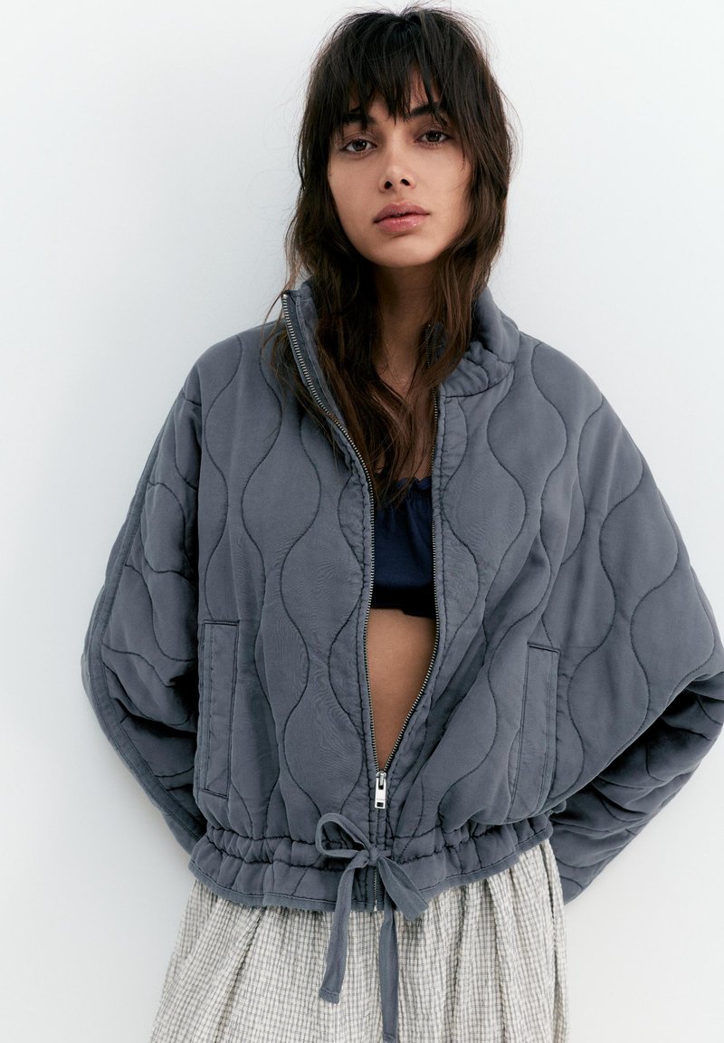 Mladá žena s vlnitými hnedými vlasmi v šedej prešívanej bunde rozopnutej cez čierne tričko a svetlú károvanú sukňu na bielom pozadí.