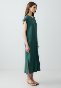 Un lungo vestito aderente in tessuto verde scuro con maniche corte, spacchi laterali e scollatura rotonda. Indossato con sandali beige.