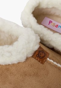 Chaussons en suede marron avec une douce doublure en fourrure crème. Présente une étiquette en cuir avec un design de singe et des coutures blanches le long du bord.