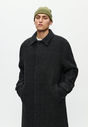Schwarz-grauer karierteter Mantel aus Wollmischung mit schickem Kragen, seitlichen Taschen und Knopfverschluss, kombiniert mit einer grünen Strickmütze.