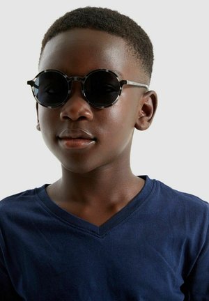 Round black sunglasses with a thin frame, worn by a person in a navy V-neck t-shirt. Smooth skin texture, short cropped hair.
