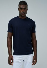 Modelo masculino vistiendo una camisa de manga corta ajustada de color azul marino y pantalones blancos, de pie frente a un fondo gris claro liso.