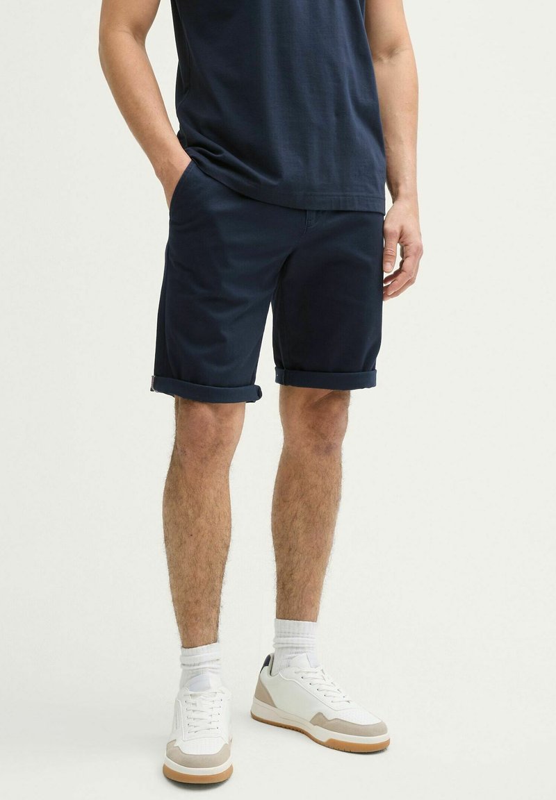 Șorturi dintr-un amestec de bumbac de culoare albastru marin, cu manșete rulate, asortate cu adidași albi și șosete albe până la gleznă. Design simplu, fără modele vizibile.