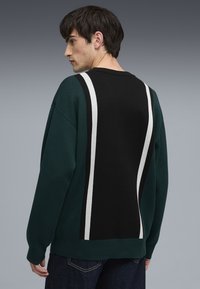 Grüner und schwarzer Pullover mit weißen Streifen auf der Rückseite, gefertigt aus einem strukturierten Strickmaterial. Lockere Passform mit abfallenden Schultern.