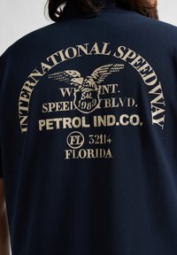 Marinblå t-shirt med vintage-inspirerade gräddfärgade grafiker med en örn och text som säger "International Speedway" och "Petrol Ind. Co."