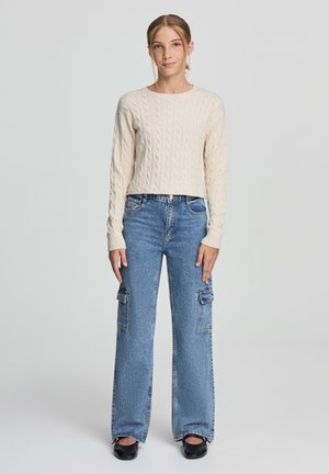 Maglione a coste in maglia crema con maniche lunghe, abbinato a jeans cargo blu a gamba larga con tasche laterali e un classico design a cinque tasche.