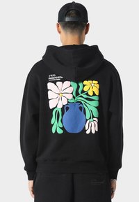 Mustat hupparit, joissa on suuri, värikäs kukkakuva, jossa on sininen maljakko, vaaleanpunaisia ja keltaisia kukkia sekä vihreitä lehtiä takana.