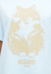 Αφράτη γάτα με στρογγυλά γυαλιά και κασκόλ, τυπωμένο σε μπεζ χρώμα σε ανοιχτό μπλε ύφασμα με το κείμενο "MOSCHINO JEANS" από κάτω.