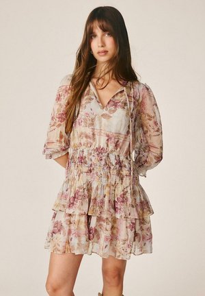 Mujer joven con cabello largo y castaño que lleva un vestido ligero de flores en capas con mangas abullonadas, de pie frente a un fondo liso.