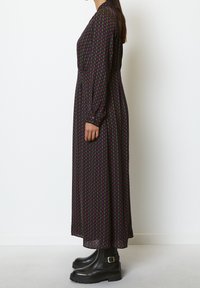 Robe maxi noire à manches longues avec un motif géométrique rose et vert. Portée avec des bottines noires à semelles épaisses, vue de côté.