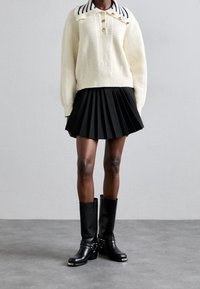 Gestrickter cremefarbener Pullover mit gestreiftem Kragen und goldenen Knöpfen, kombiniert mit einem schwarzen plissierten Rock und schwarzen kniehohen Stiefeln mit metallischen Akzenten.