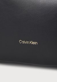 Čierna kožená taška s hladkým povrchom; embosované logo "Calvin Klein" v zlatej farbe. Jednoduchý, minimalistický dizajn bez ďalších kovových prvkov.