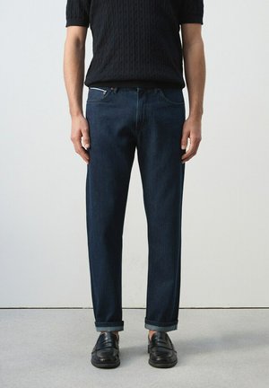 Hombre con vaqueros azul oscuro con puños doblados, mocasines negros y una camisa de punto negra de manga corta, de pie contra un fondo liso.