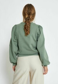 Green sweatshirt with puffed long sleeves and a fitted waist. Fabric appears soft and slightly textured, paired with light beige trousers.