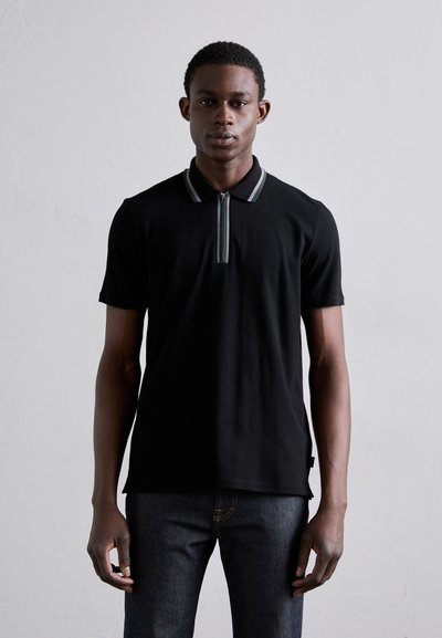 Polo noir à manches courtes, col zippé avec des accents verts et gris, associé à un jean en denim foncé. Texture du tissu lisse.
