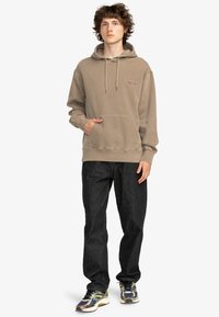 Brun bomullshuva med framficka och dragsnören, kombinerad med svarta jeans och flerfärgade sneakers, visad på en stående manlig modell.