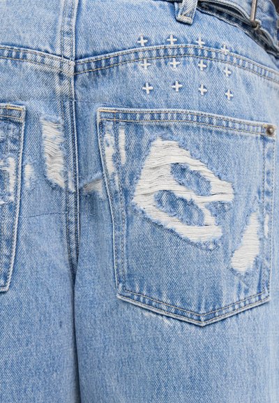 Jeans de mezclilla decolorados en azul claro que presentan parches deshilachados y acentos de punto cruz cerca de los bolsillos traseros. Cierre de botón metálico visible.