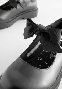 Chaussures Mary Jane en cuir noir avec une semelle plate, ornées d'un accent en forme de nœud texturé et d'une doublure noire à motifs d'étoiles.
