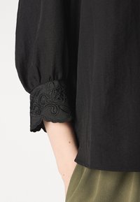 Blusa nera con maniche a sbuffo arricciate e pizzo nero ricamato decorativo al polsino. Il tessuto ha una consistenza liscia.