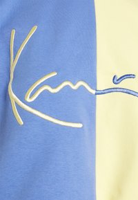 Košile s krátkým rukávem s modrožlutými barevnými bloky, s vyšitým textem v krémové a světle modré barvě, což dodává texturu a detail.