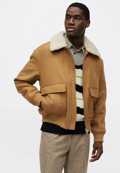 Καφέ μάλλινο σακάκι με μπεζ ψεύτικη γούνα στον γιακά, διαθέτει δύο μπροστινές τσέπες, κλείσιμο με φερμουάρ και ριμπ μανσέτες, φορεμένο πάνω από πουκάμισο με σχέδιο.