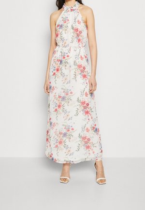 Kobieta ubrana w białą, bezrękawnikową sukienkę maxi z różowo-fioletowym kwiatowym wzorem i falbaniastego dekoltu na szyję, w połączeniu z białymi butami na otwartym palcu.
