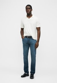 Moški stoji z eno roko v žepu, oblečen v belo polo majico s kratkimi rokavi, modre kavbojke, črni pas in črne čevlje, na enobarvnem ozadju.