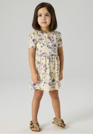 Fille aux cheveux bruns mi-longs portant une robe crème à manches courtes avec un imprimé floral violet et marron, ainsi que des sandales beige à boucle.