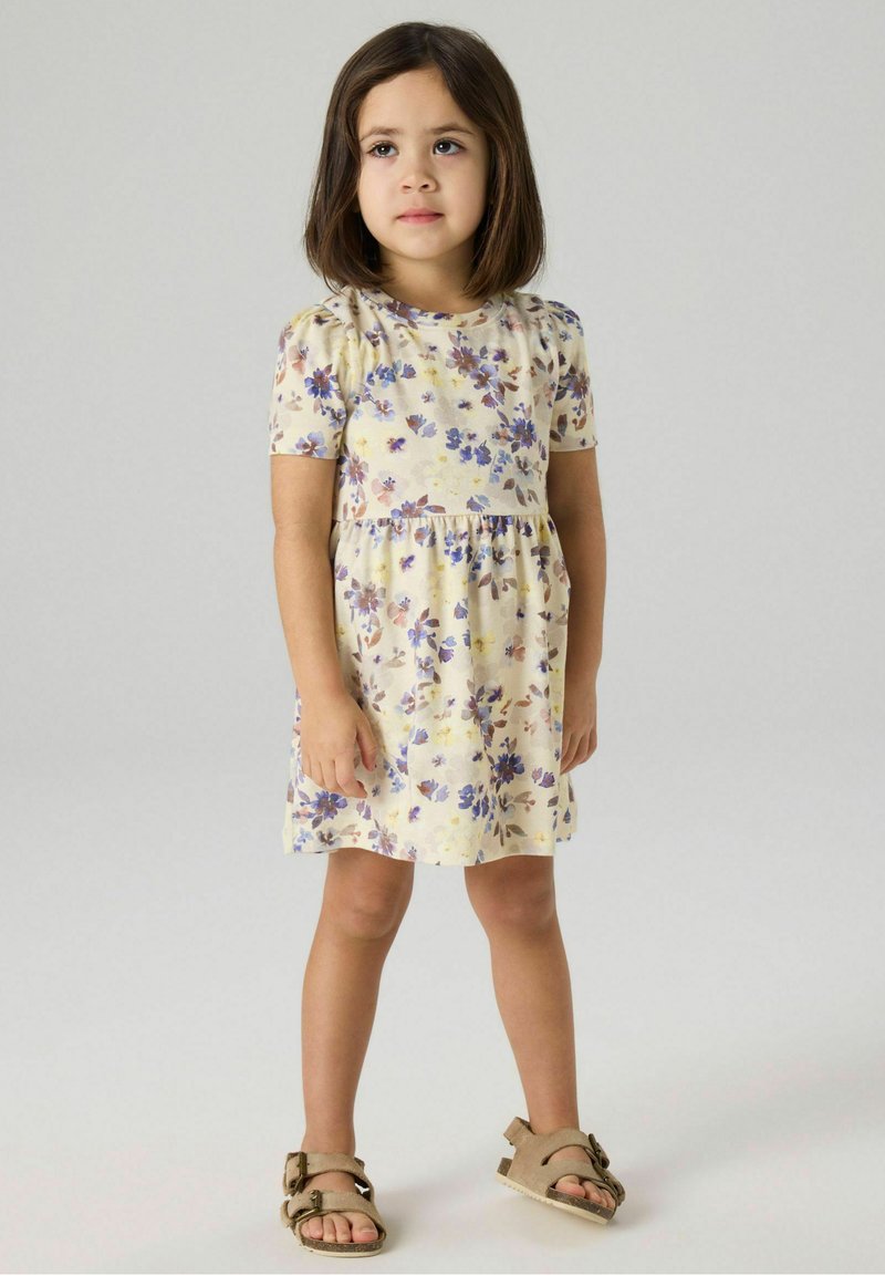 Κορίτσι με σκούρα μαλλιά μέχρι τους ώμους φοράει κοντομάνικο κρεμ φόρεμα με μοτίβο λουλουδιών σε μωβ και καφέ αποχρώσεις και μπεζ σανδάλια με αγκράφα.