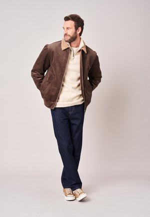Man die een bruine ribcord jas draagt, een crèmekleurige trui, donkere spijkerbroek en beige sneakers, staat met zijn handen in de zakken en kijkt opzij.