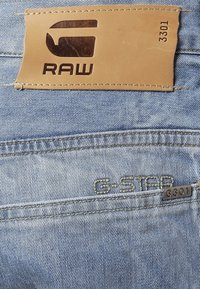 Jeans de mezclilla azul claro con parche de cuero que dice "RAW", "G-STAR" bordado y una etiqueta metálica "3301" en el bolsillo trasero.