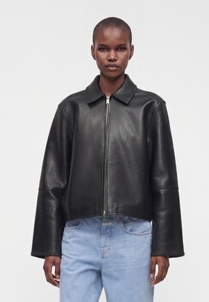 Persona che indossa una giacca di pelle nera con zip e maniche larghe e jeans azzurri su uno sfondo chiaro uniforme.