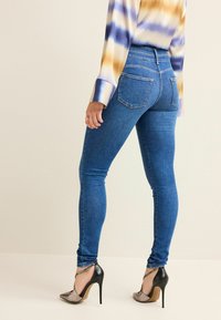 Jeans skinny bleus avec une texture lisse, taille haute et deux poches arrière. Portés avec des chaussures à talons hauts noires et un haut à motif.