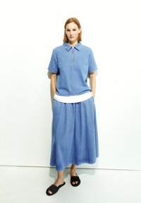 Denim-Polohemd mit kurzen Ärmeln, ausgestattet mit einem Reißverschluss, kombiniert mit einem langen, fließenden Denim-Rock. Schwarze Rutschsandalen runden das Outfit ab.