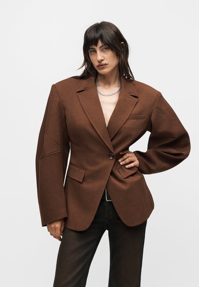 Άτομο με σκούρα μαλλιά που φοράει ένα υπερμεγέθες καφέ σακάκι και μαύρο παντελόνι, ποζάροντας με το ένα χέρι στη μέση μπροστά σε απλό φόντο.