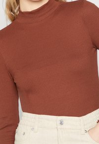 Brauner, gerippter Stehkragen-Top kombiniert mit hellen, elastischen Hosen mit elastischem Bund, die strukturierten Stoff und einfache Designdetails zeigen.