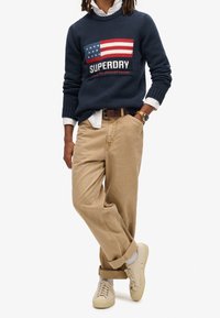 Pull marine avec motif du drapeau américain, pantalon en velours côtelé beige et baskets beiges. Détails remarquables : texture tricotée et revers aux ourlets du pantalon.