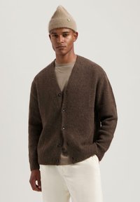 Bruin cardigan met een gestructureerde afwerking, voorzien van een V-hals, knoopsluiting en lange mouwen, gelaagd over een lichtbeige shirt.