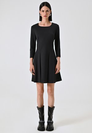 Schwarzes A-Linien-Kleid mit drei Viertelärmeln, runder Halslinie, plissiertem Rock, kombiniert mit knielangen schwarzen Stiefeln mit Nietenakzenten.