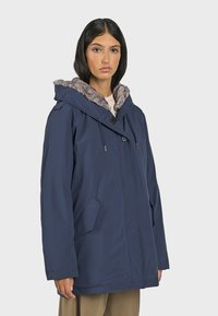 Marineblauwe parka met een nepbont-gevoerde capuchon, voorkeursluiting, twee zijzakken en een verstelbare trekkoord in de capuchon.