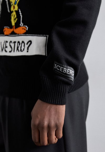Le pull en tricot noir présente un design de personnage de bande dessinée, des poignets côtelés et une écusson de marque sur la manche. Texture lisse et coupe ajustée.