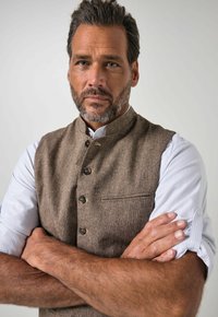Hombre con los brazos cruzados, lleva un chaleco marrón sobre una camisa blanca con las mangas remangadas, mirando directamente con una expresión seria.