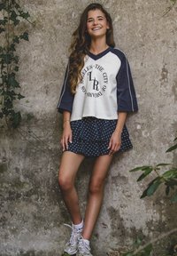 Witte en marineblauwe sporttop met grafische print, gecombineerd met een marineblauwe gestapelde rok met polkadots. Model draagt witte sneakers en enkelsokken.