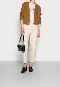 Braune gerippte Strickjacke, cremefarbenes T-Shirt mit Aufdruck, beige Hose, schwarze Slipper und eine schwarze Handtasche mit Kettenakzenten.