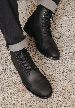 Black leather boots with a rounded toe, lace-up design, and smooth texture. Cuffed gray pants reveal a contrasting fabric detail at the hem.