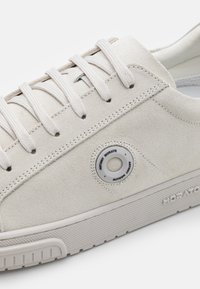Világos bézs velúr sneaker kerek fűzőlyuk részlettel, bézs fűzőkkel és texturált gumitalppal. Egyszerű dizájn, kontrasztos varrási díszítéssel.
