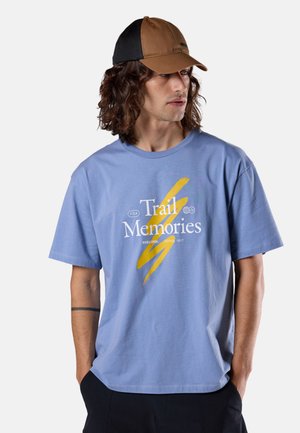 Mężczyzna o kręconych włosach do ramion nosi brązowo-czarną czapkę, niebieską koszulkę z napisem „Trail Memories” i czarne spodnie, ma ręce w kieszeniach, patrzy w prawo.