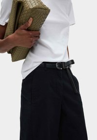 Zwarte wijde broek met zijzakken, gecombineerd met een strakke witte t-shirt. Een gestructureerde olijfgroene geweven clutch wordt in de hand gehouden.