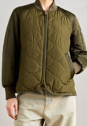 Personne portant une veste bomber matelassée vert olive avec fermeture éclair devant et un pantalon beige clair, main dans la poche de la veste.