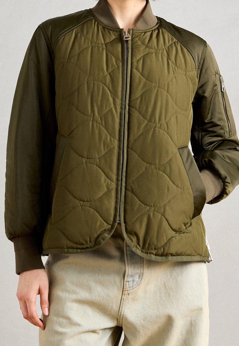 Personne portant une veste bomber matelassée vert olive avec fermeture éclair devant et un pantalon beige clair, main dans la poche de la veste.